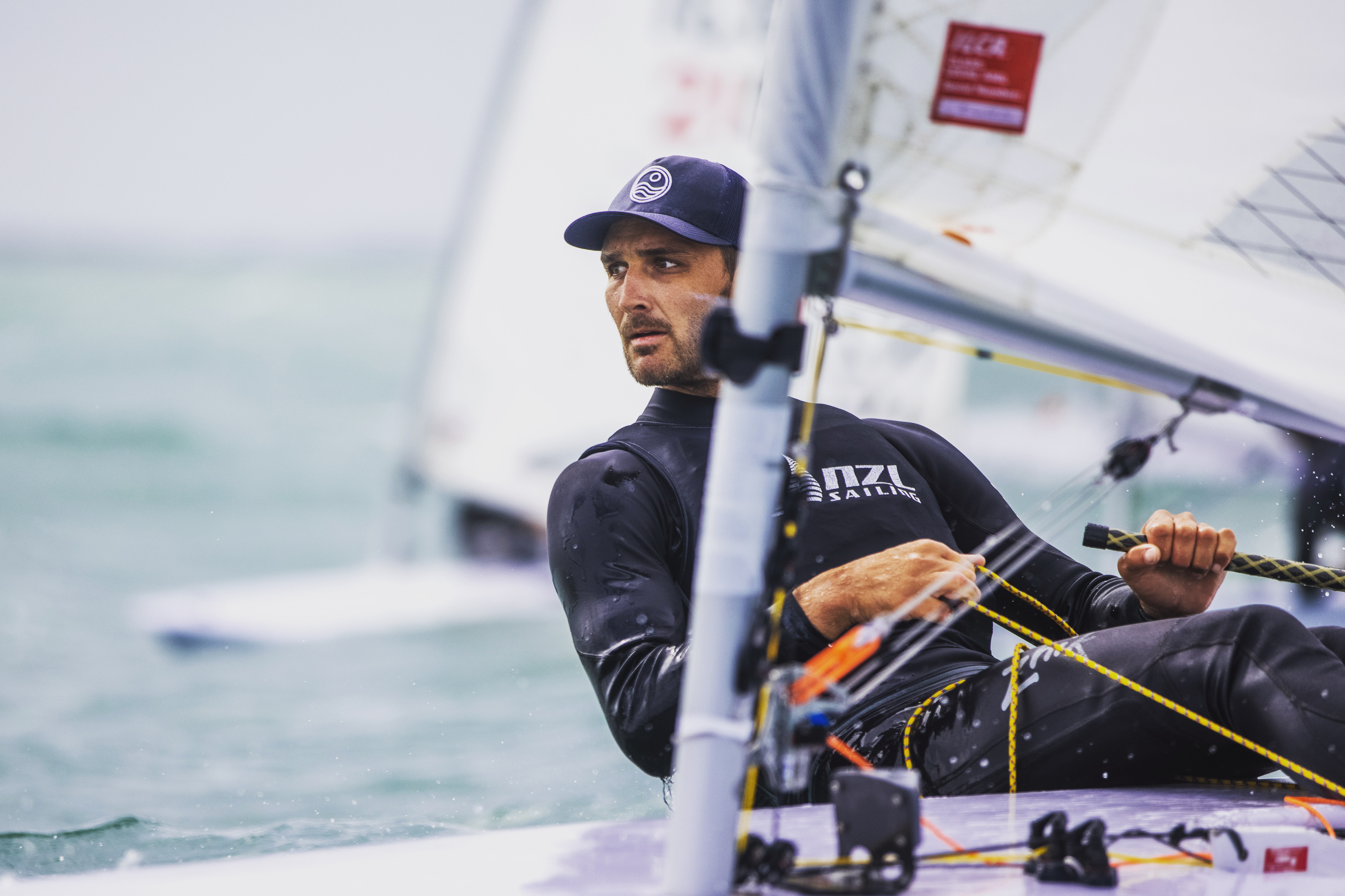 A man in a black wetsuit and baseball cap, holding a sailboat pole, appears to be participating in a sailing competition. ...