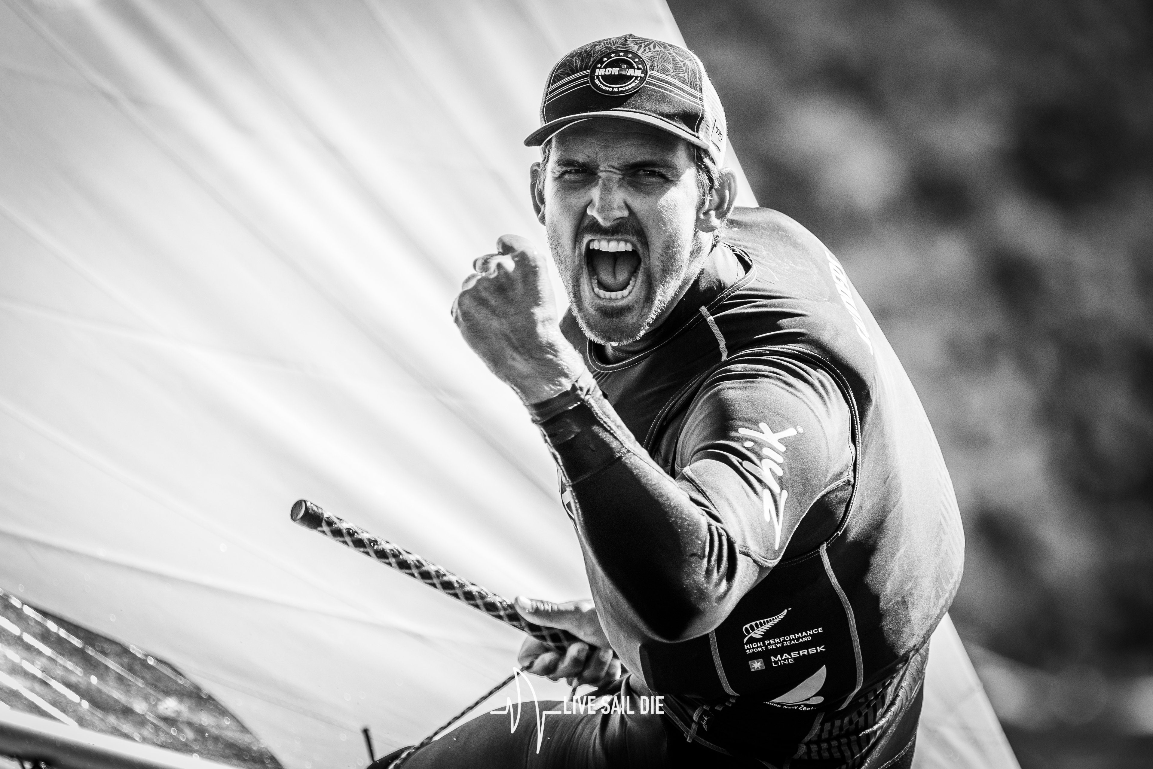 A man in a baseball cap and long-sleeved shirt with a black and white logo on the chest, holding a boat's steering wheel a...