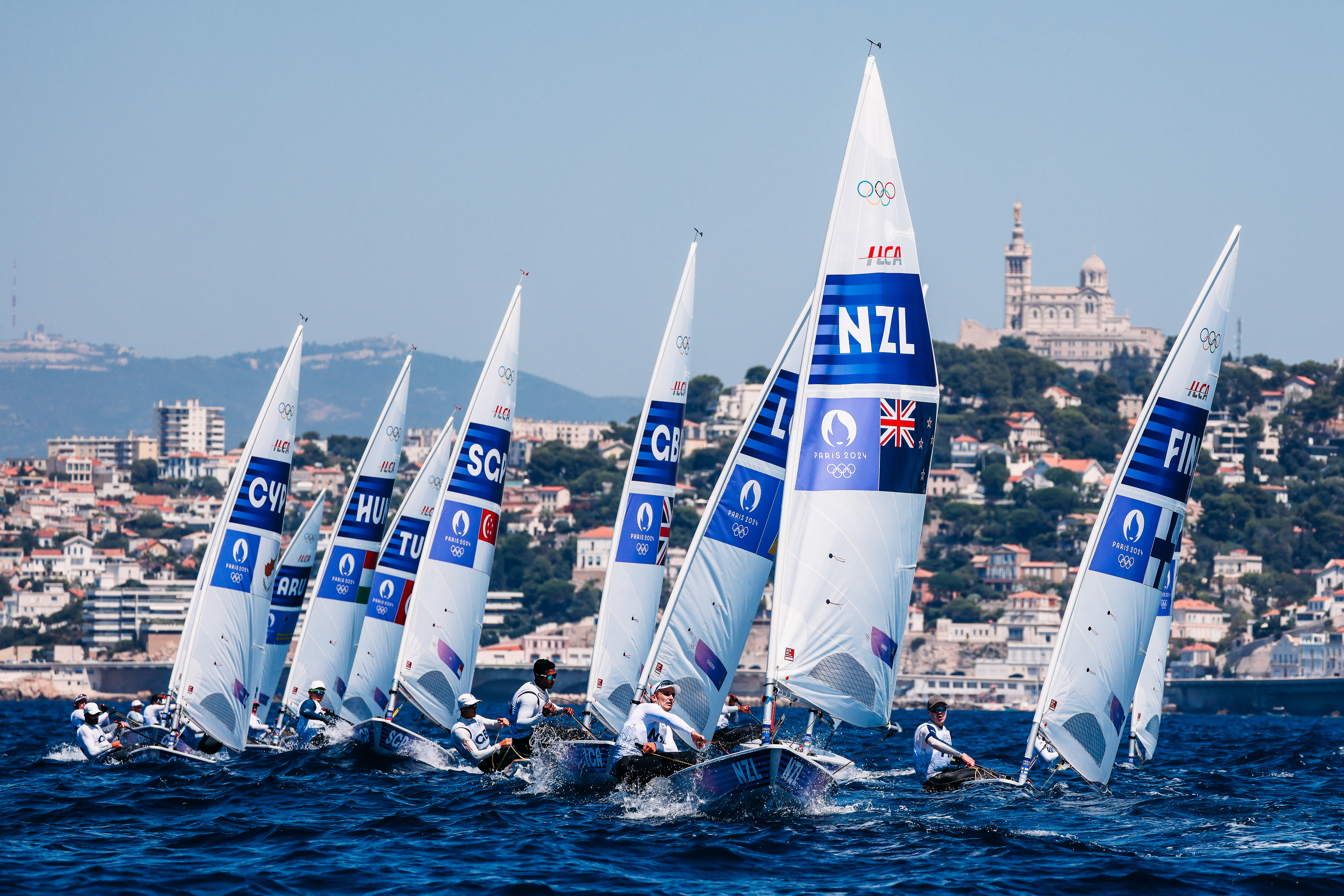 A fleet of sailboats with white sails and blue logos, featuring the Olympic rings and the letters NZL, race across a large...
